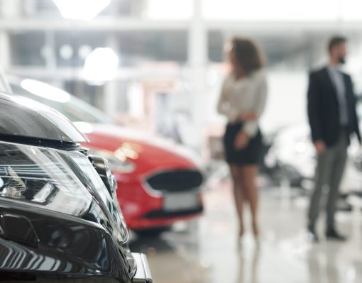 Close up of black car headlight. Modern showroom with expensive automobiles. People standing in car center, looking for and choosing vehicles. Woman and man observing auto for purchase.